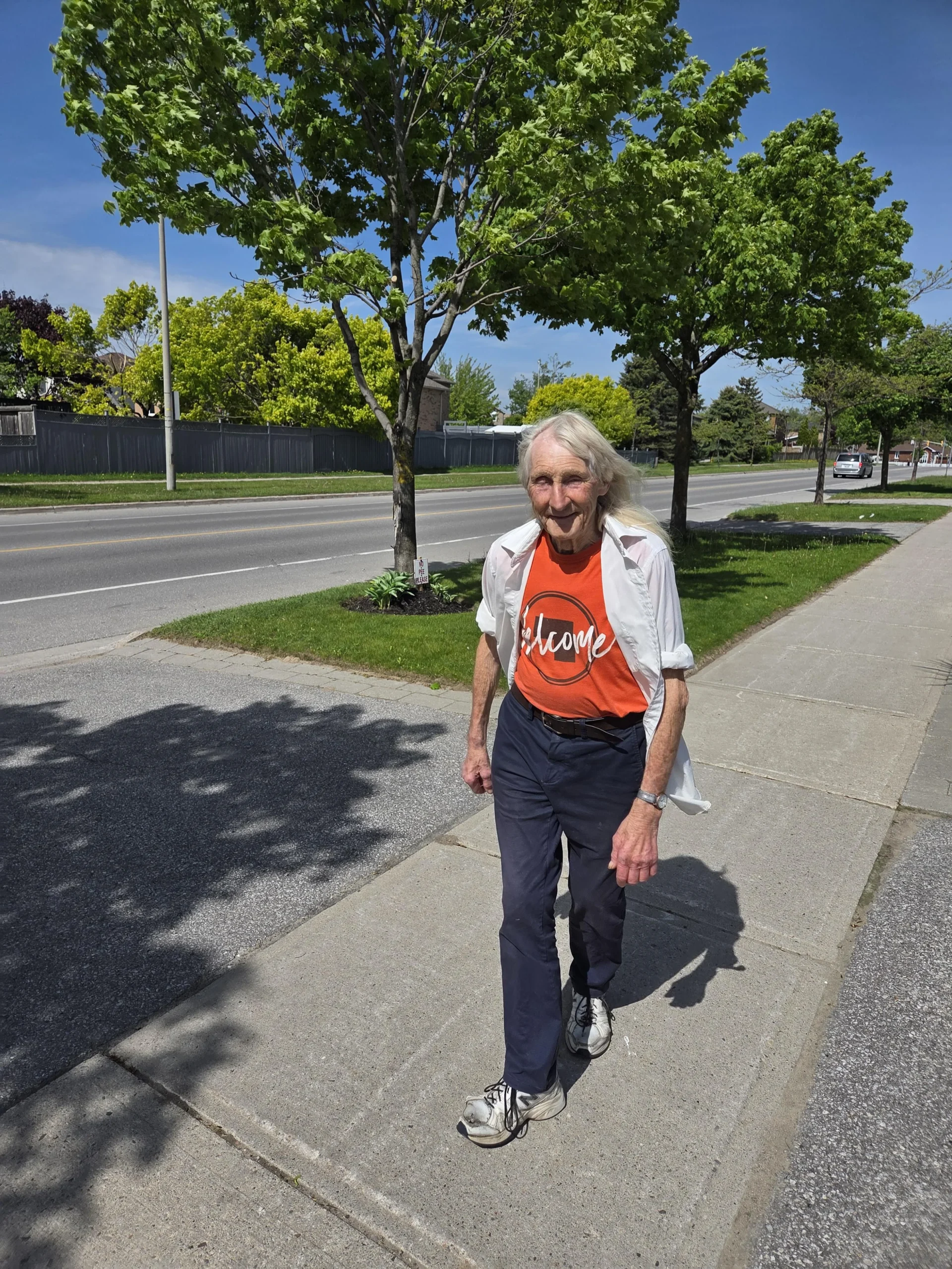 An older person with long white hair walks on a sunny sidewalk, wearing a red "Welcome" shirt, white open button-down, navy pants, and sneakers. Trees line the street and the sky is clear and blue.