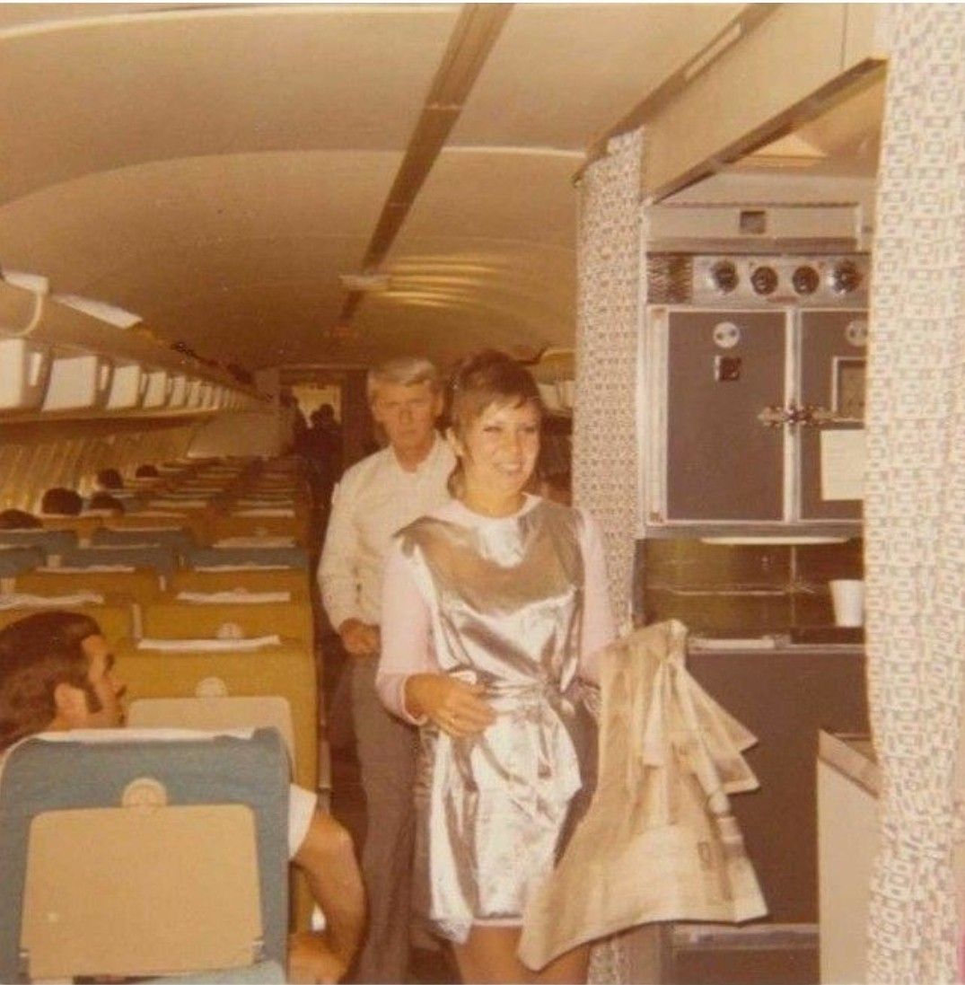 A smiling flight attendant in a shiny silver uniform stands near the airplane galley, holding a folded blanket, with passengers seated and one man walking down the aisle in the background.
