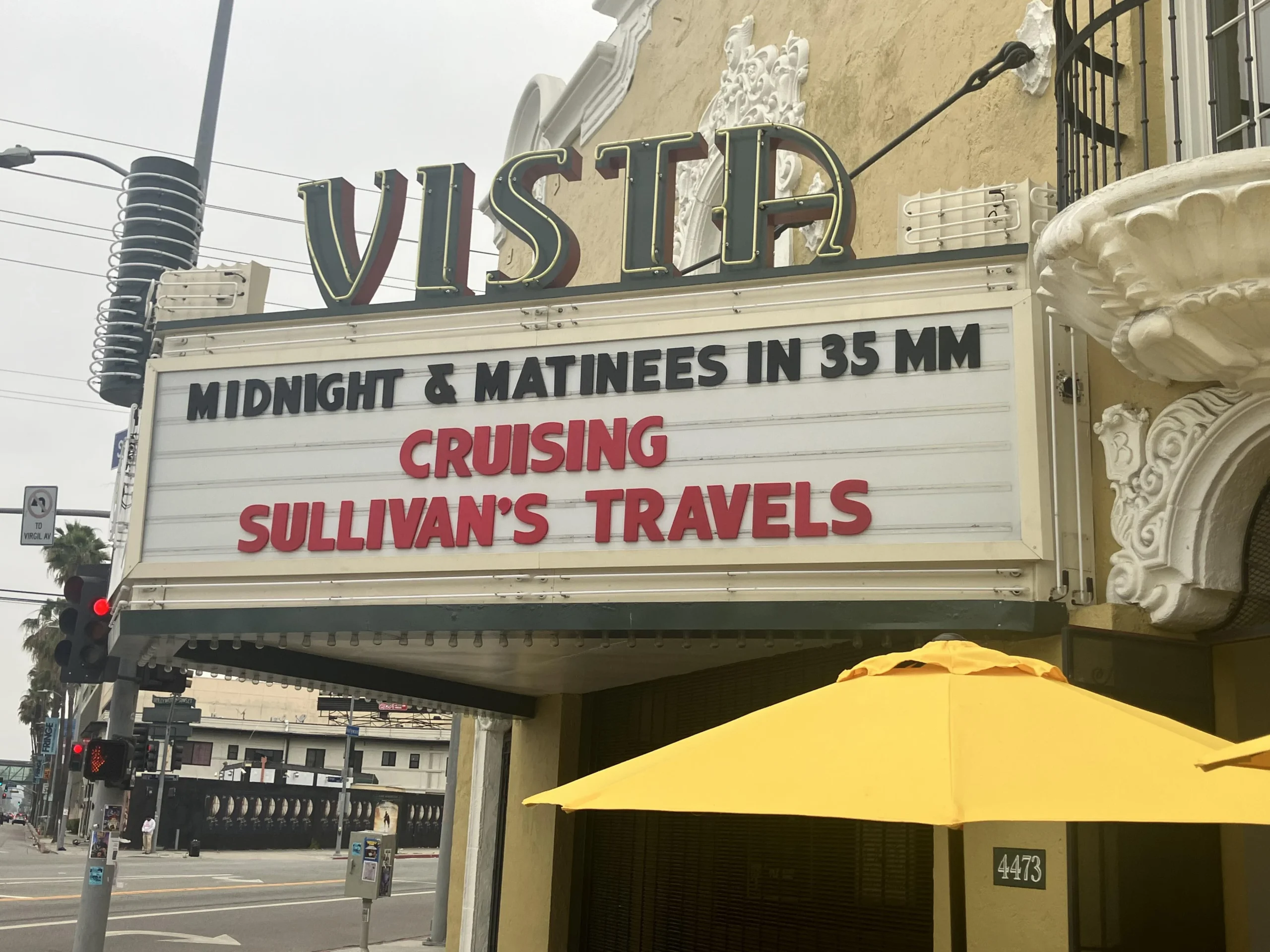 The Vista Theater marquee displays: “MIDNIGHT & MATINEES IN 35 MM CRUISING SULLIVAN’S TRAVELS.” A yellow umbrella and street intersection are visible in front of the building.