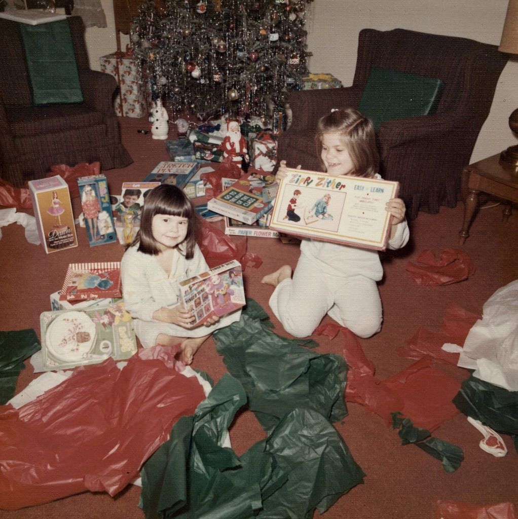 Two young children in pajamas sit on the floor amid torn wrapping paper, holding up toys and books, with a decorated Christmas tree and presents in the background.