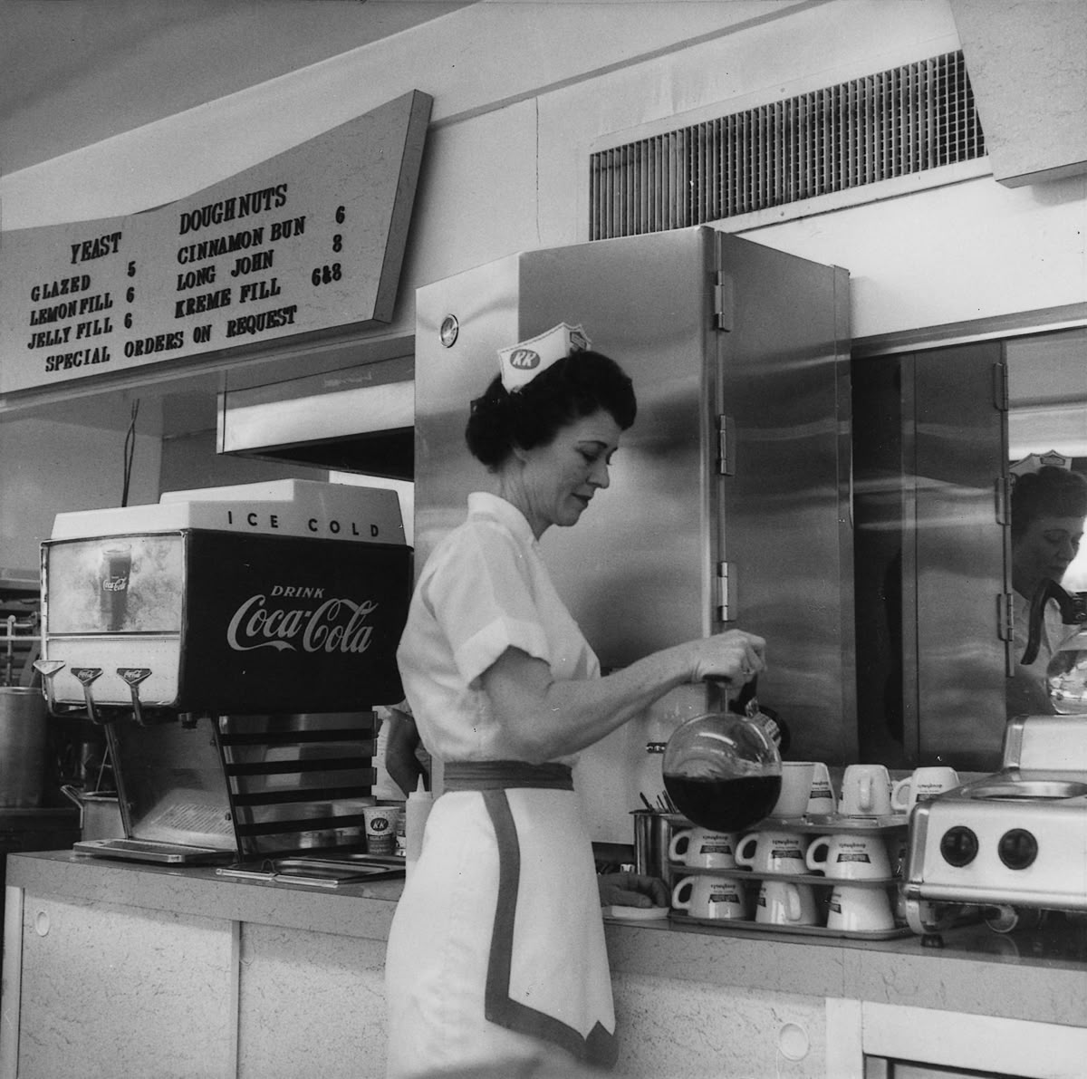 A waitress in a retro diner uniform pours coffee behind the counter, with a Coca-Cola cooler, stacked mugs, and a menu board with doughnut options visible in the background.