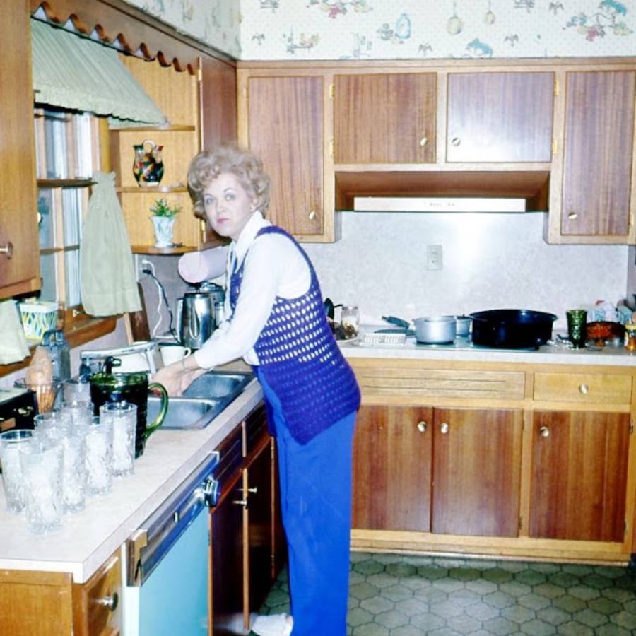 A woman in a blue vest and white shirt stands at a kitchen sink, washing dishes. The kitchen has wooden cabinets, glassware on the counter, and pots on the stove. She looks toward the camera.