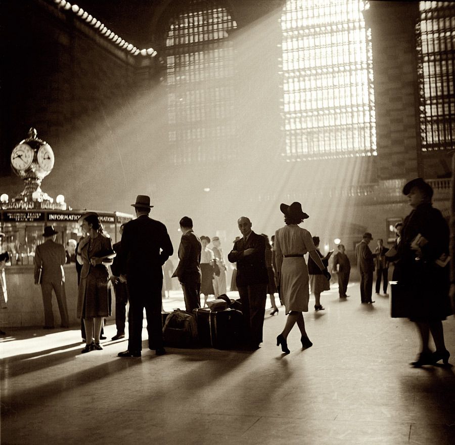 Sepia-toned photo of people in vintage clothing standing in a grand train station lobby with sunlight streaming through tall windows, illuminating the crowd and a clock above an information booth.