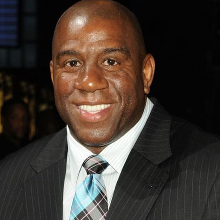 A man in a pinstripe suit with a blue striped tie smiles at the camera against a dark background.