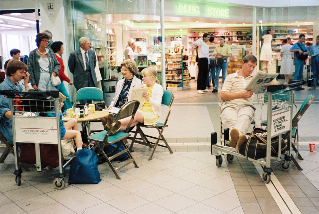 People sit on chairs and luggage carts in an airport terminal near a drug store; some read newspapers, others talk, and a few children sit with adults. Luggage sits nearby on carts. The scene looks busy yet relaxed.