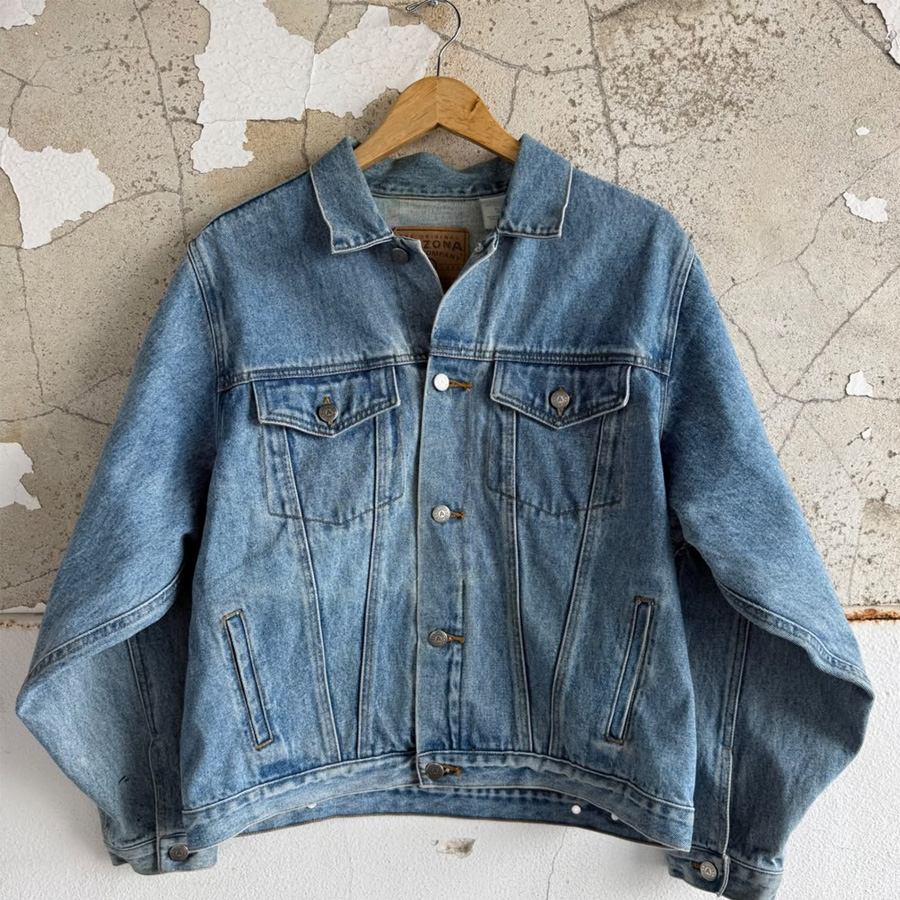 A blue denim jacket hangs on a wooden hanger against a textured, cracked white and beige wall. The jacket has buttoned chest pockets and silver buttons down the front.