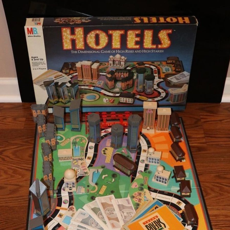 A board game called "Hotels" is set up, showing a colorful game board with miniature hotels, play money, cards, and the game box in the background.
