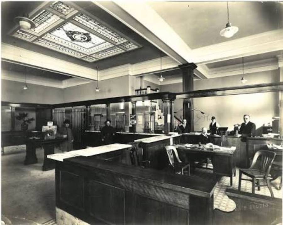 Black-and-white photo of a vintage bank interior with wooden counters, marble surfaces, and a decorative ceiling. Several people, both seated and standing, are working or assisting customers. The atmosphere is formal and orderly.
