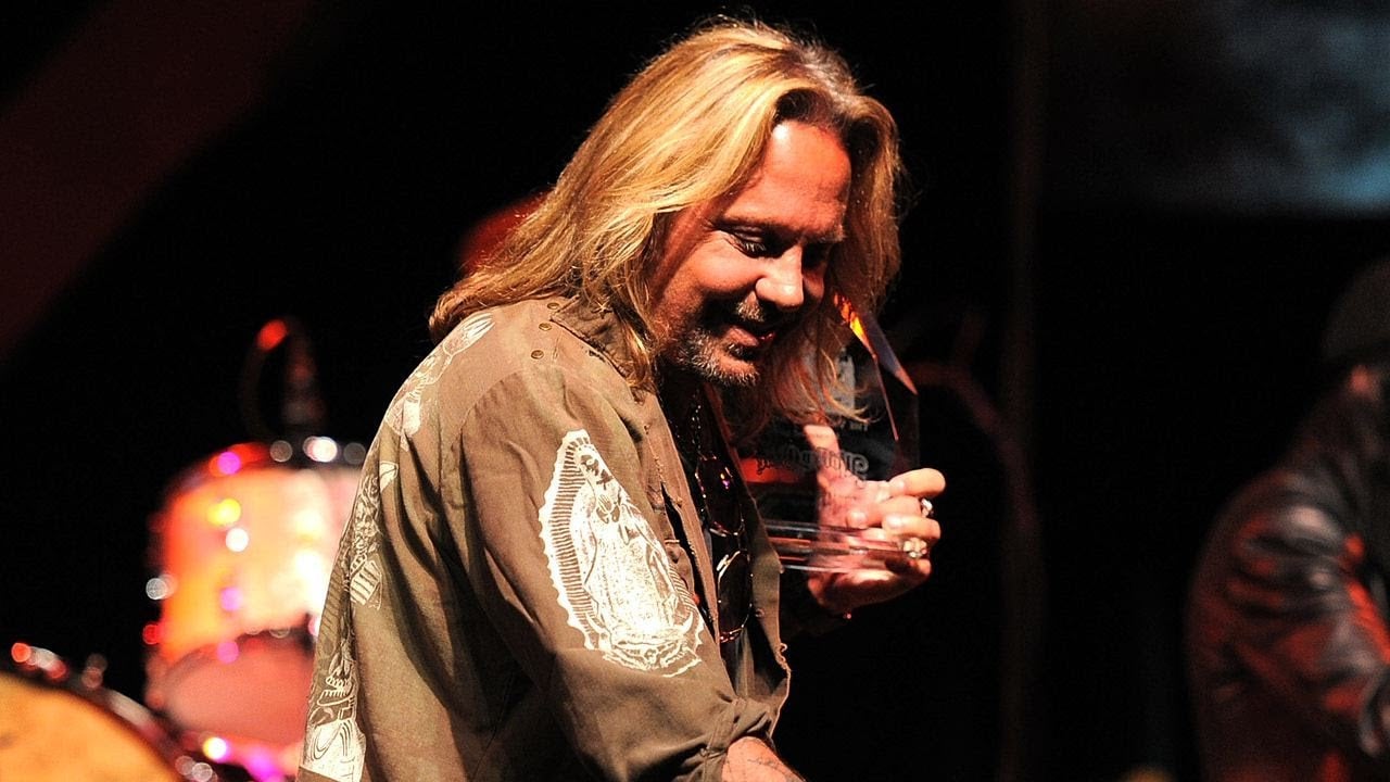 A man with long blonde hair, wearing an embroidered shirt, holds a glass award on stage with drums and musical equipment in the background.