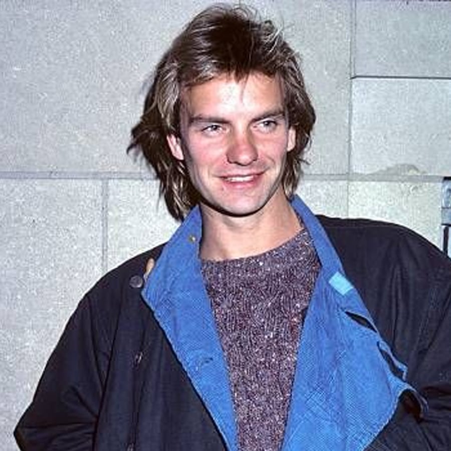A man with light brown, shaggy hair and a relaxed smile wears a textured sweater and dark jacket with a blue lining, standing in front of a stone wall.