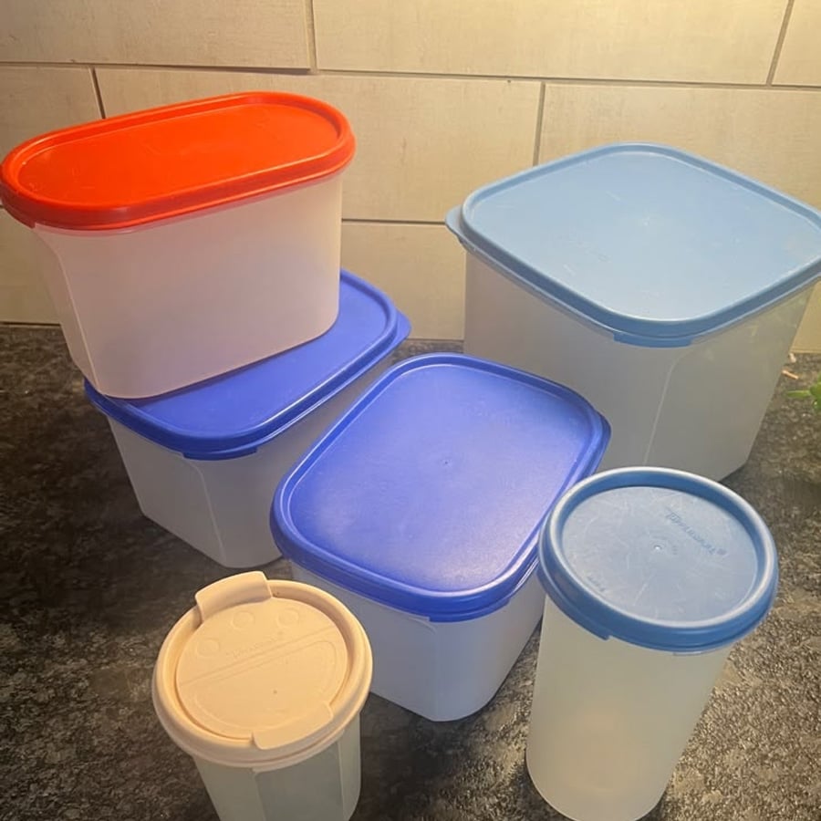 Six plastic containers with blue, red, and off-white lids of different shapes and sizes are arranged on a dark countertop against a tile wall.