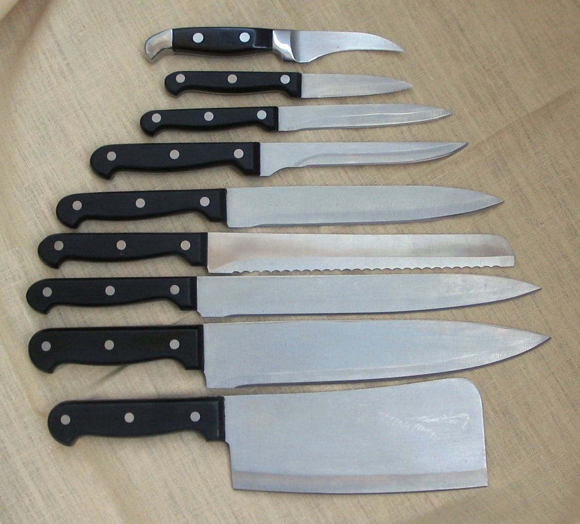 Eight kitchen knives of various sizes and shapes with black handles are arranged in a row on a beige fabric surface. The blades vary, including a serrated one and a cleaver at the bottom.