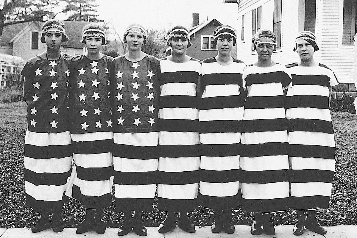 Seven women stand in a row outdoors, wearing dresses that resemble the American flag, with stars on the top and horizontal stripes on the skirts, likely in a patriotic or historical context.