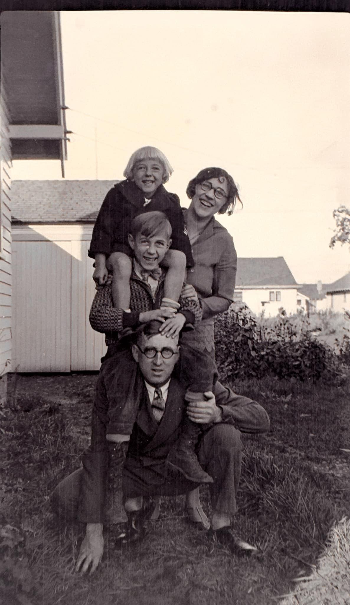 A black-and-white photo shows a smiling family stacked playfully outdoors. An older man kneels on the ground, with a boy sitting on his shoulders, a woman standing behind, and a young girl perched on top, all looking happy.