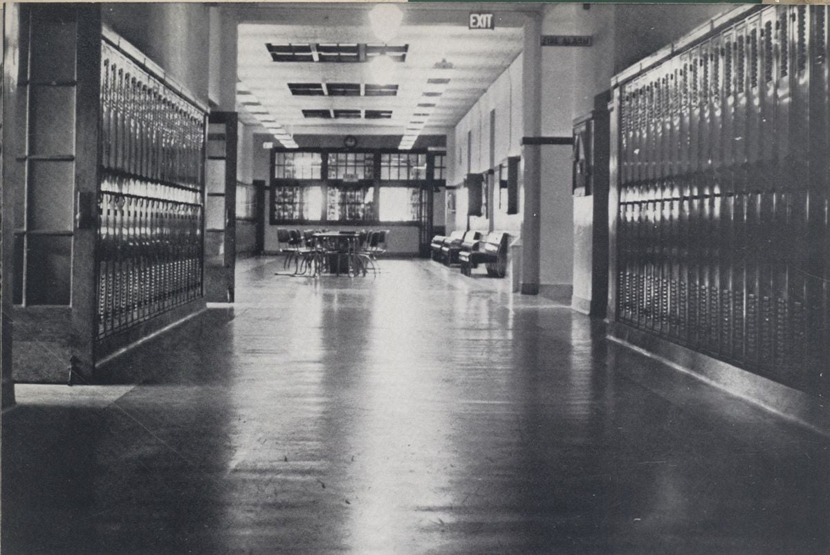 Black and white photo of an empty school hallway with rows of lockers on both sides, a few chairs and couches in the distance, and an illuminated exit sign on the ceiling.
