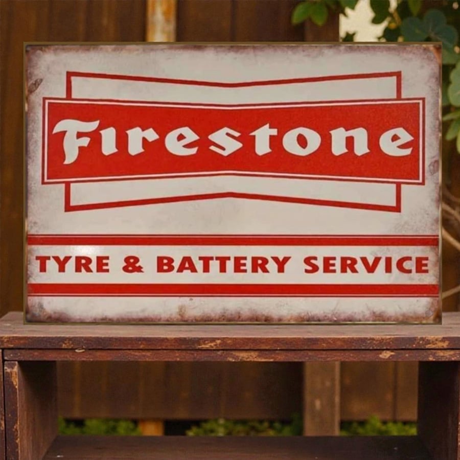 A vintage metal sign shows the Firestone logo and reads "Tyre & Battery Service" in red and white lettering, placed on a wooden surface outdoors.
