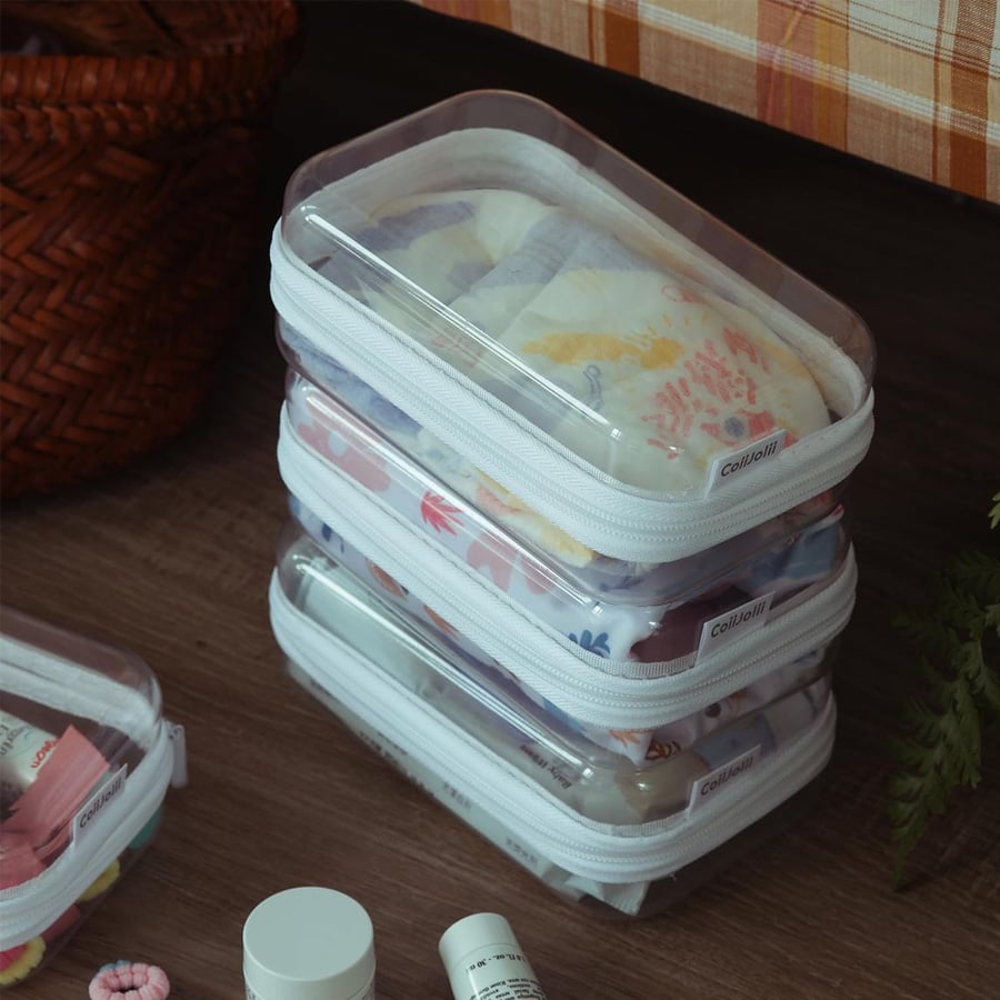 Three clear zippered pouches stacked on a wooden floor contain baby diapers with colorful prints. A woven basket and bottles of skincare products are nearby. The setting appears to be a cozy, organized room.