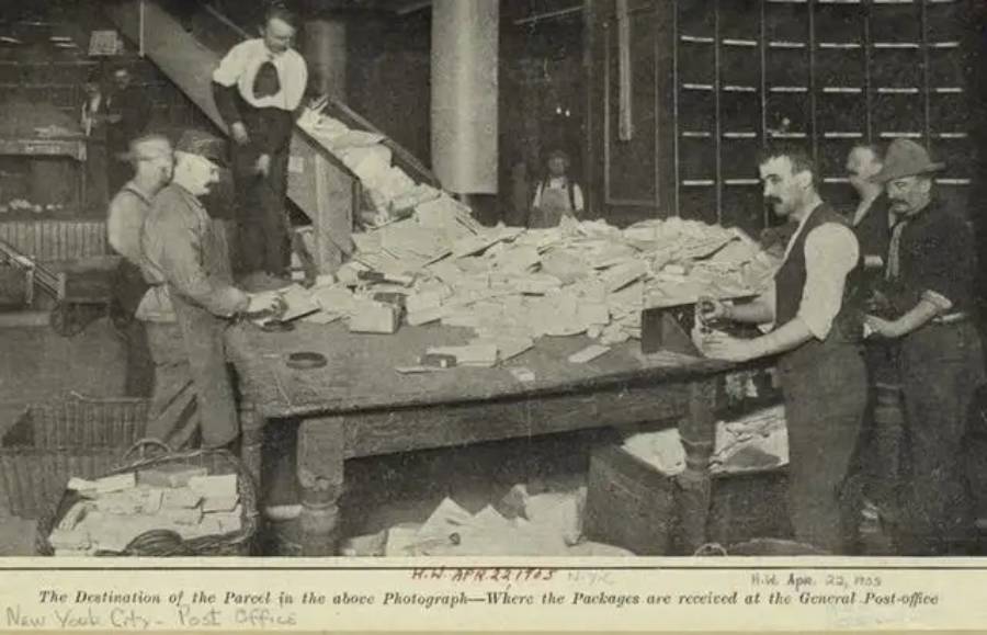 Four men sort large piles of mail and packages on a long table inside a busy post office, with shelves and sacks of mail in the background. The scene appears old-fashioned, likely from the early 20th century.