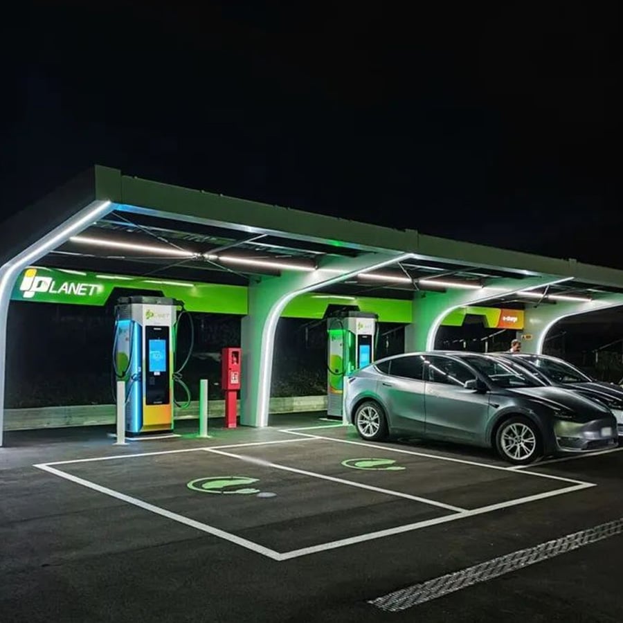 Two electric cars parked at night under a well-lit, modern EV charging station canopy labeled "PLANET," with glowing green accents and multiple charging stalls.