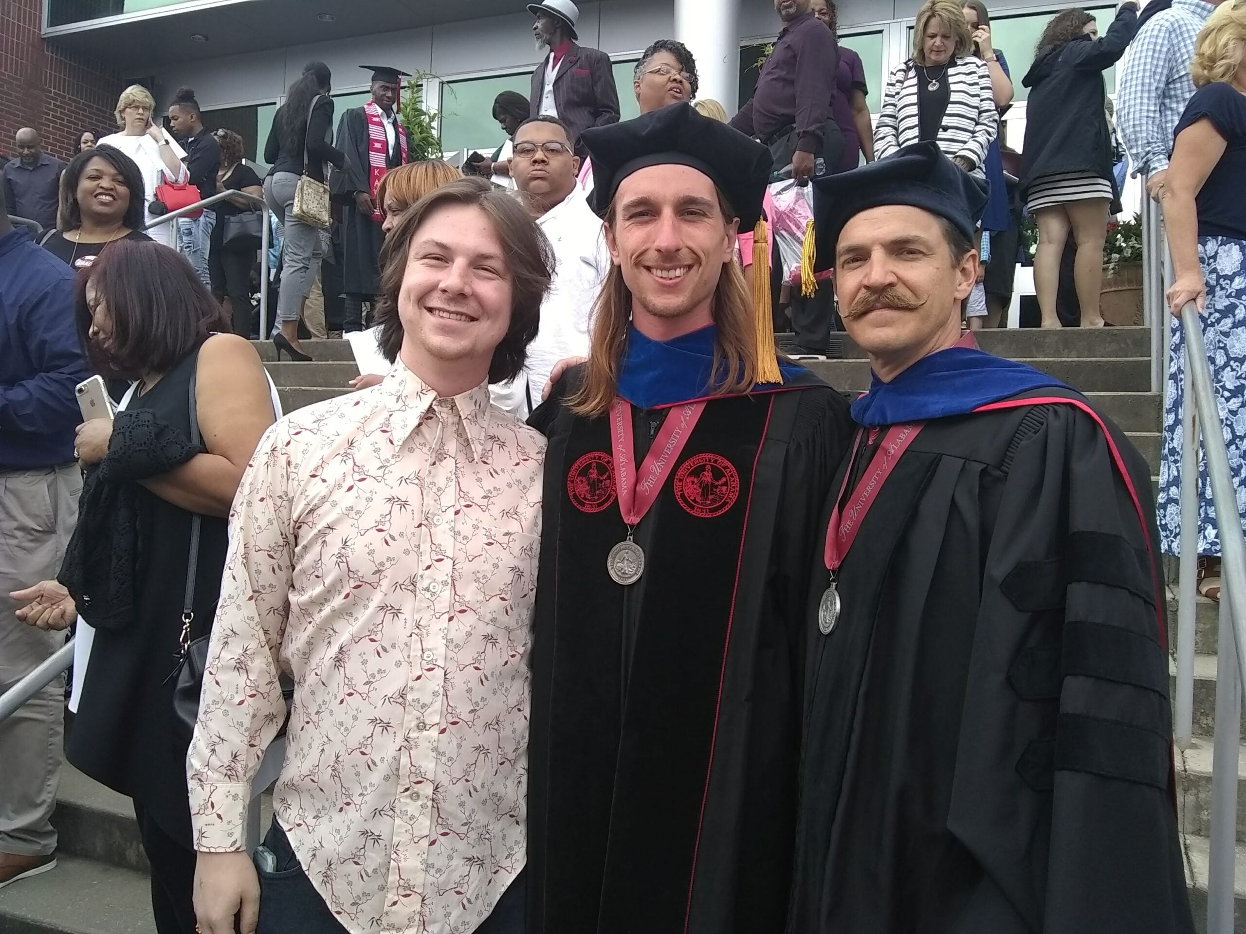 Three smiling people pose outside at a graduation ceremony. Two wear academic robes and caps with medals, while the third person wears a patterned shirt. A crowd stands on steps in the background.