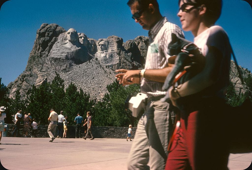 Tourists walk in front of Mount Rushmore on a sunny day, with the monument’s four presidential faces carved into the mountain visible in the background. Some people stand and admire the view, while trees line the area.