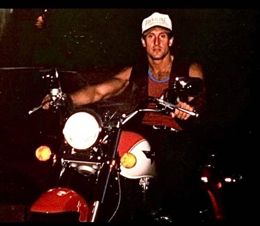 A man wearing a sleeveless shirt and a white cap sits on a red and white motorcycle at night, looking directly at the camera.