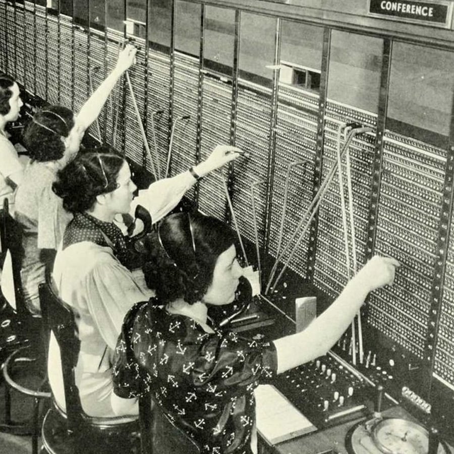 Four women sit at an old-fashioned telephone switchboard, wearing headsets and connecting calls by plugging in cables. The workspace is filled with rows of jacks, switches, and labeled "CONFERENCE" above.