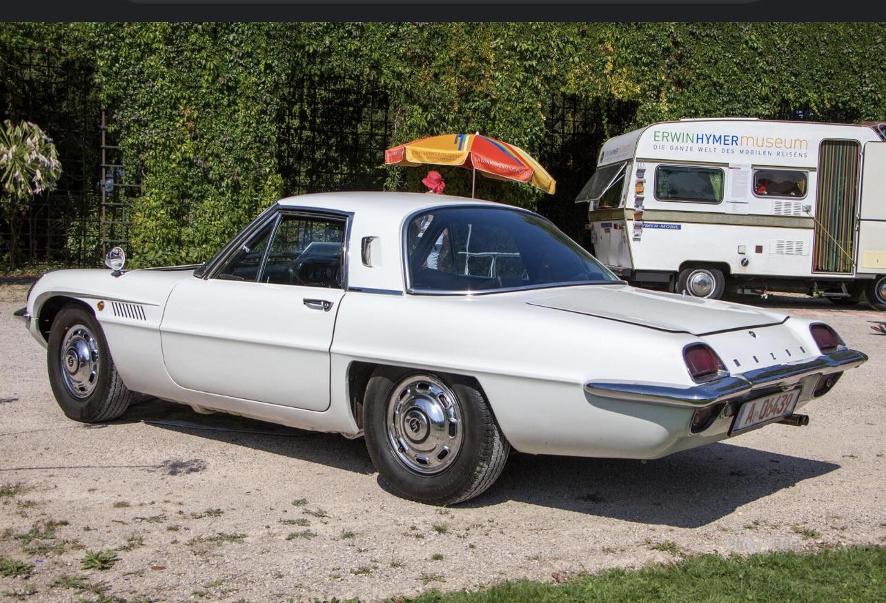 A classic white sports car parked on gravel near a food truck and a colorful umbrella, with trees and greenery in the background. The car has vintage styling and chrome wheels.