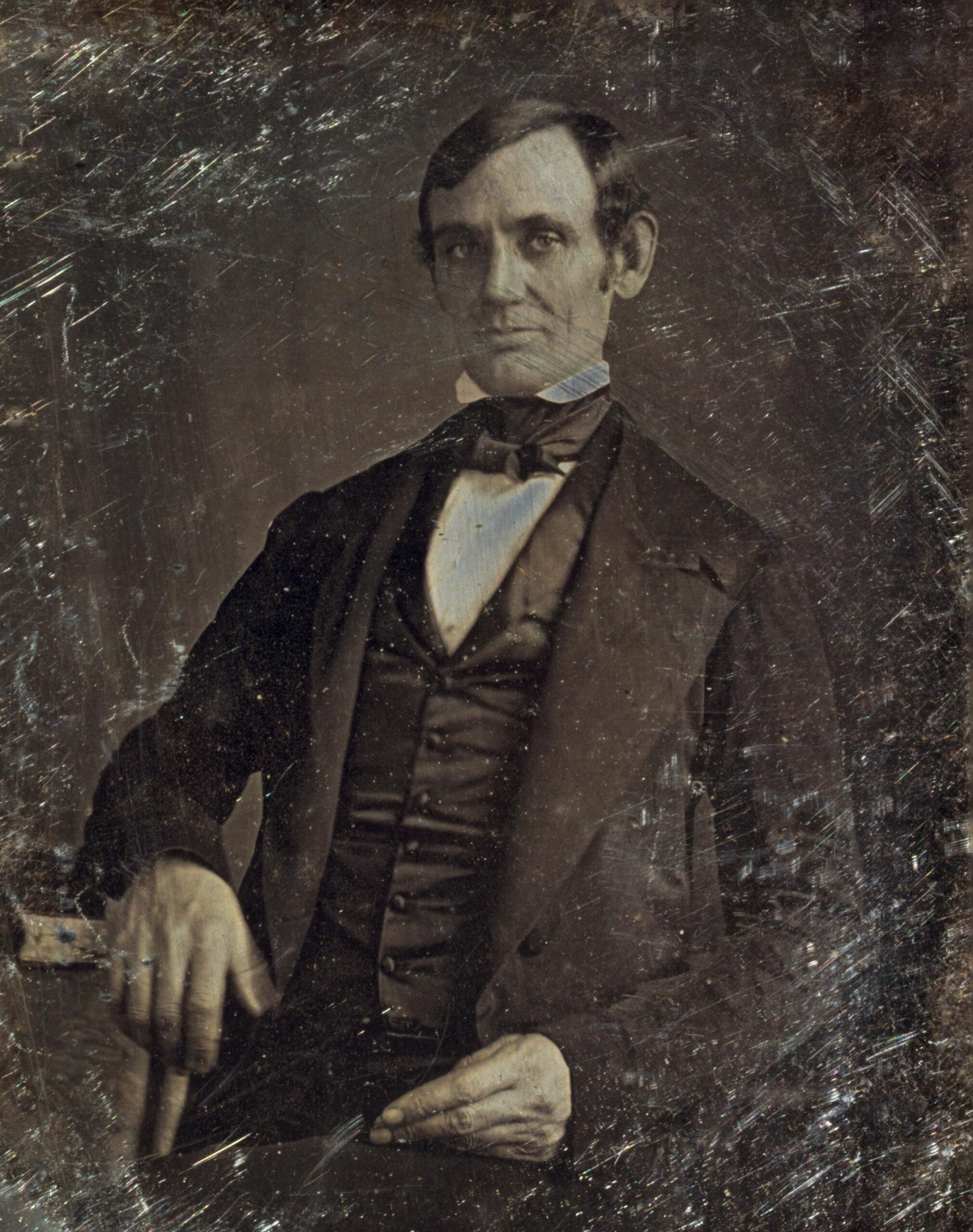 A seated man in a suit with a bow tie, looking slightly to the side. The photograph is old and has visible scratches and signs of wear.