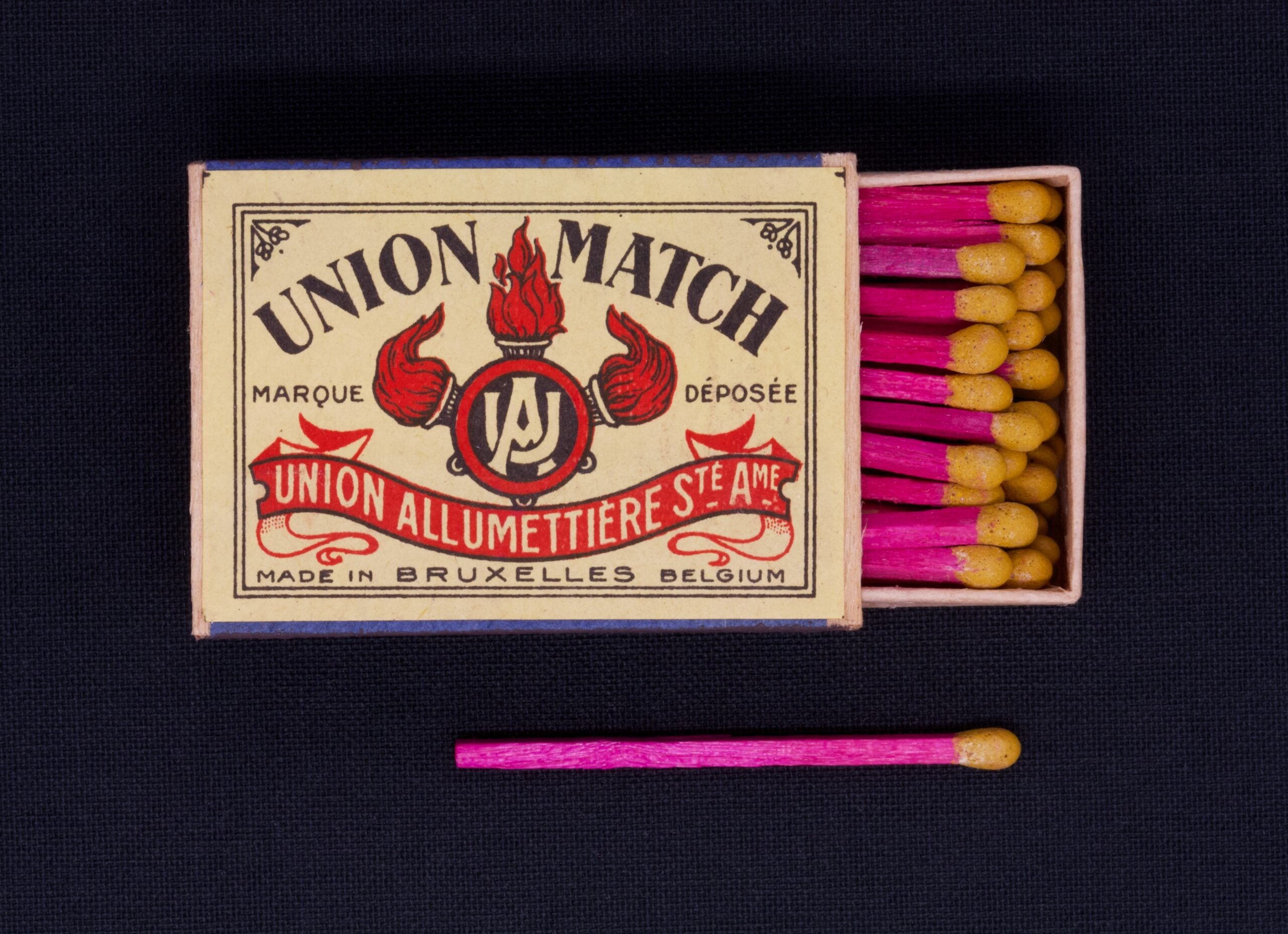 A vintage Union Match box with pink-tipped matches, one match lying outside the open box. The label reads "Union Allumettière Ste Ame, Made in Bruxelles Belgium.