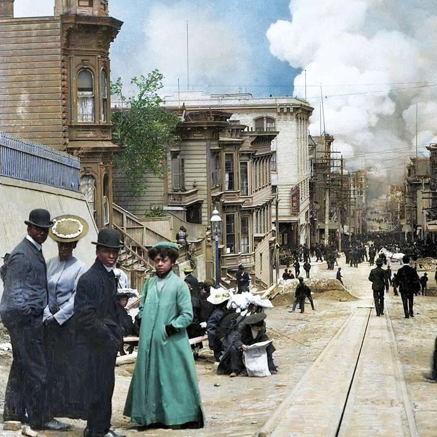 People in early 1900s attire stand on a street with damaged buildings and smoke in the background, likely after a disaster. Debris and workers fill the road.