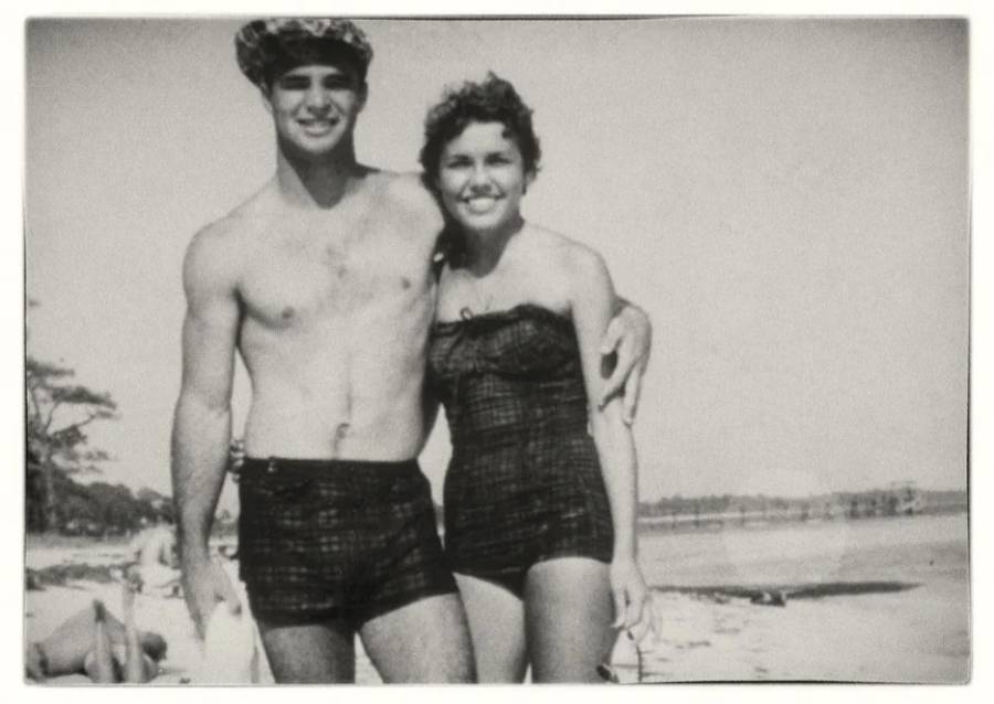 A young man and woman in vintage swimsuits stand arm-in-arm and smiling on a sandy beach, with trees and water in the background.