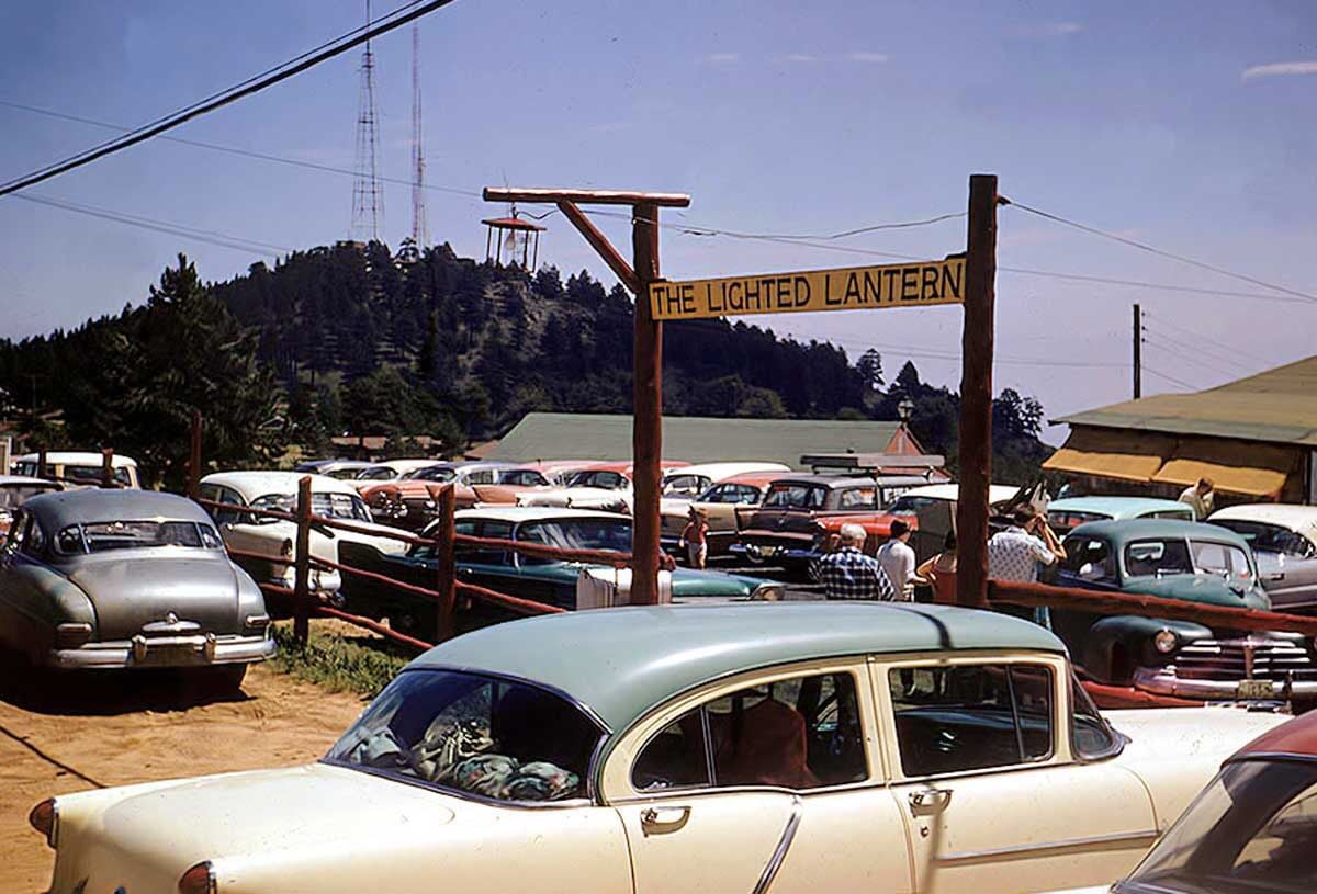 A parking lot filled with vintage cars sits beside a wooden fence. A sign reading "The Lighted Lantern" hangs in the foreground. Trees, hills, and radio towers are visible in the background under a clear sky.
