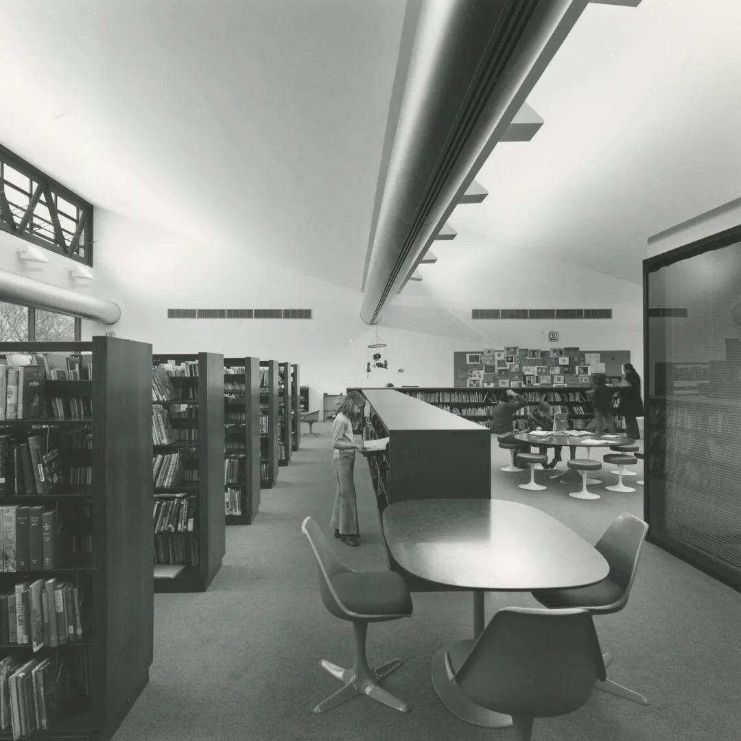 Black and white photo of a modern library interior with bookshelves, chairs, tables, and a few people reading or browsing books. The space features high ceilings, large windows, and sleek, contemporary furniture.