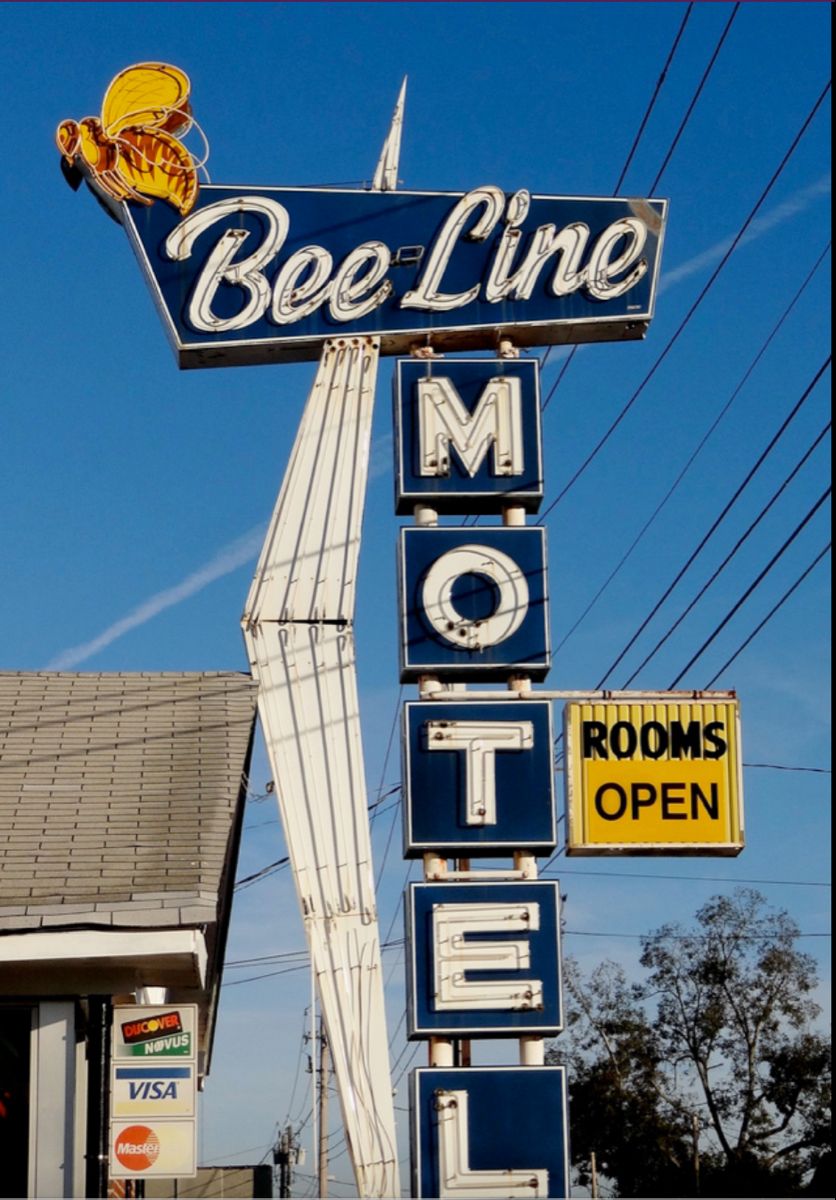 Vintage roadside sign reads “Bee-Line MOTEL” with a bee graphic. A yellow sign says “ROOMS OPEN.” The sign is against a blue sky, with wires and part of a building visible.