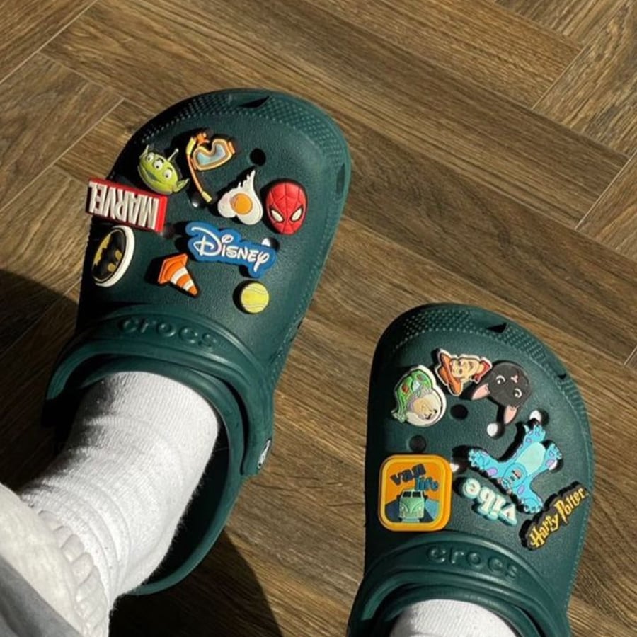A person wearing white socks and green Crocs decorated with colorful character charms from Disney, Marvel, Harry Potter, and Stitch, standing on a wooden floor.