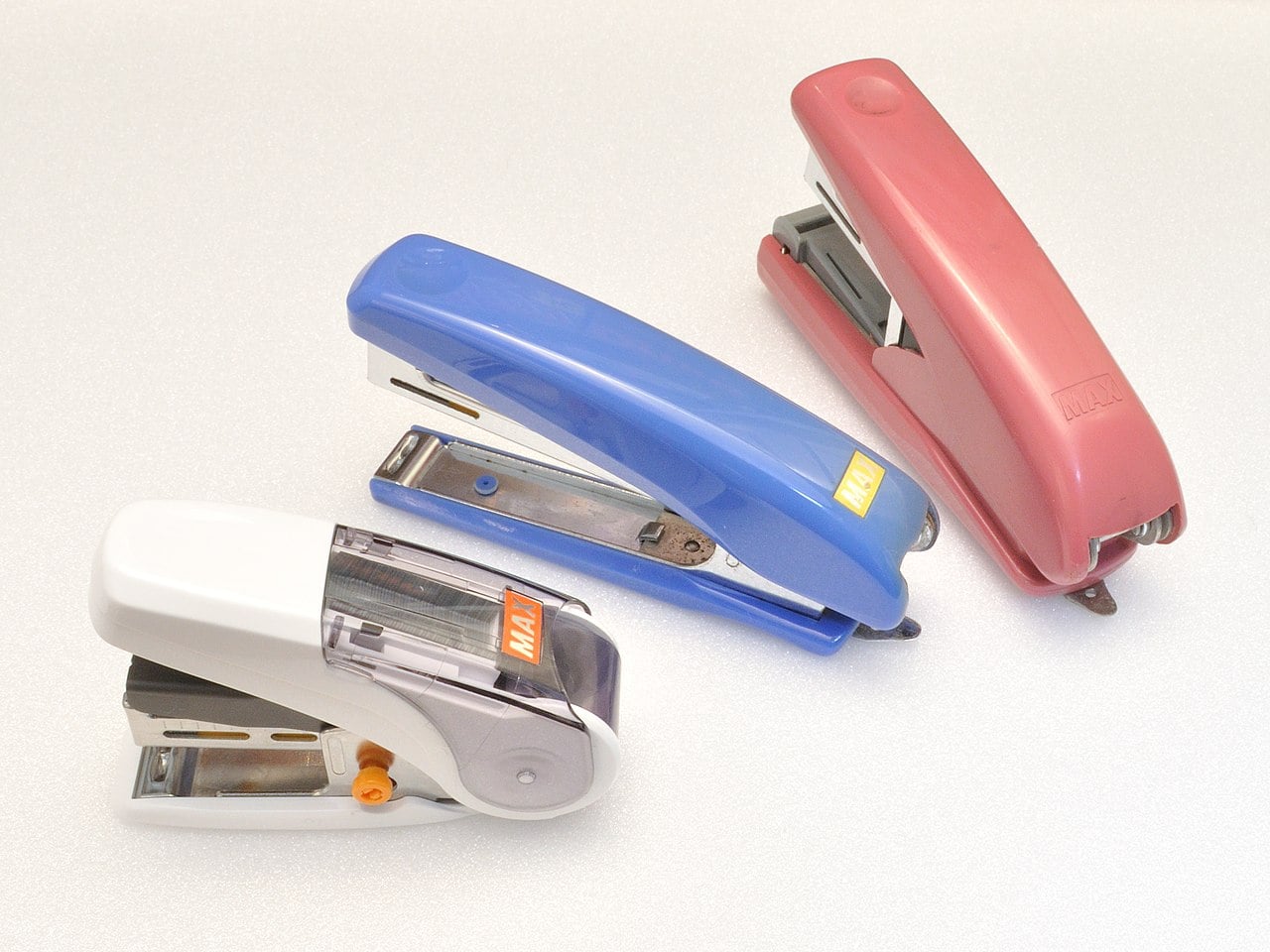 Three handheld staplers—one white, one blue, and one red—are arranged on a light surface. The white and blue staplers are partly open, while the red stapler is closed.