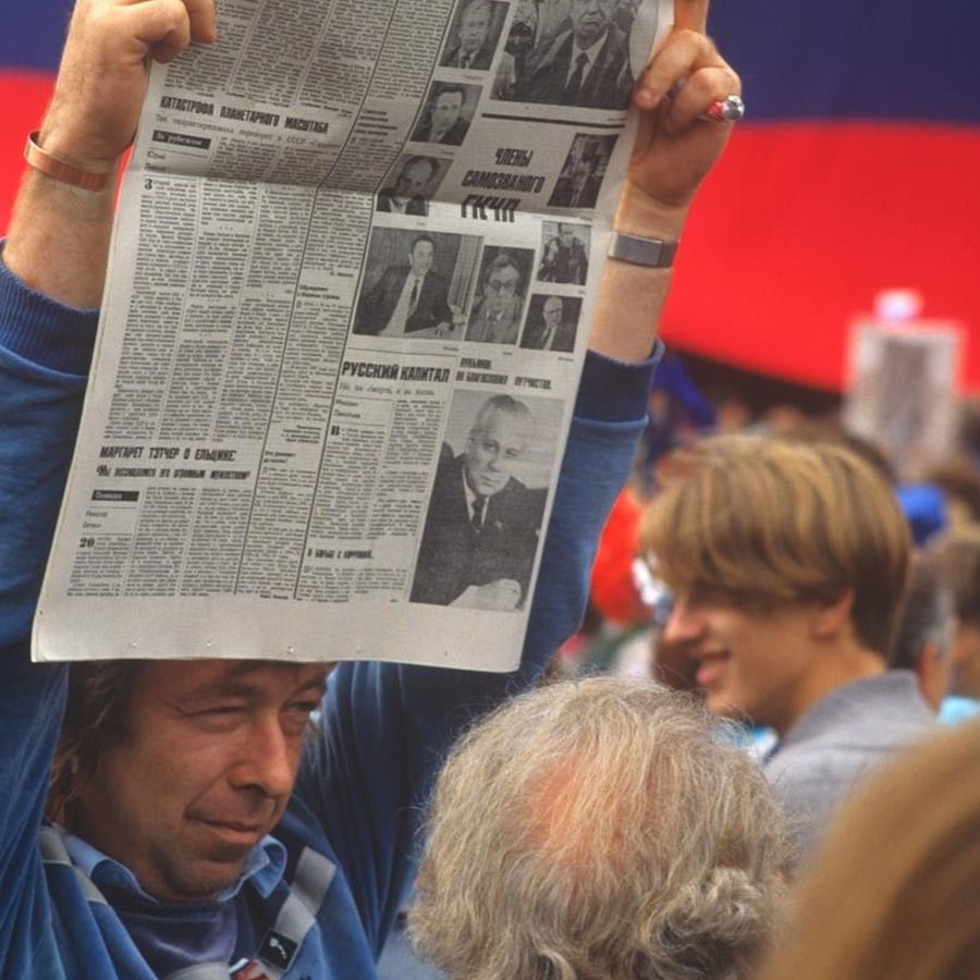 Person holds up a Russian-language newspaper in a crowd; several people are visible in the background, and a large red and blue flag is partially visible behind them.