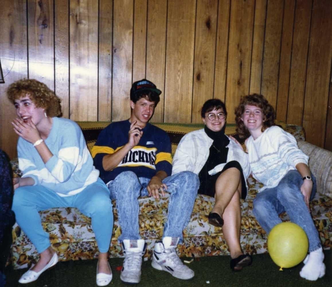 Four young adults sit on a couch against wood-paneled walls. They are smiling and laughing. One holds up a peace sign. A yellow balloon is on the floor in front of them. The setting appears casual and retro.