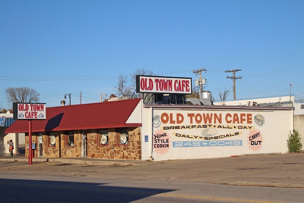 A small diner with a red roof and stone exterior has signs reading “Old Town Cafe” and advertises breakfast all day, daily specials, homestyle cooking, and carry out service. The sky is clear and sunny.