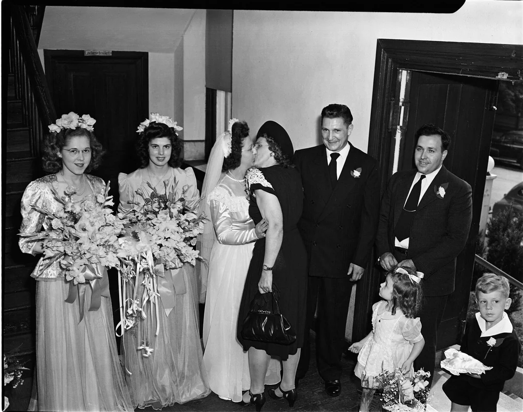 A bride in a white dress is kissed on the cheek by a woman in black, surrounded by bridesmaids, men in suits, a young girl, and a boy with flowers, all indoors on a wedding day.