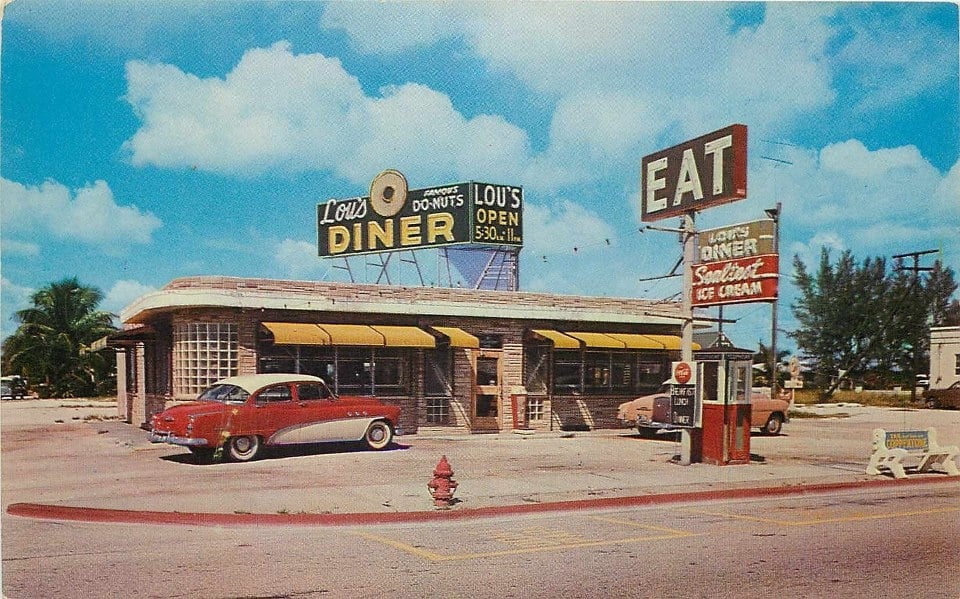A vintage diner with yellow awnings and retro signs, including one reading "EAT." Classic cars are parked outside, and palm trees are visible in the background under a blue sky with scattered clouds.