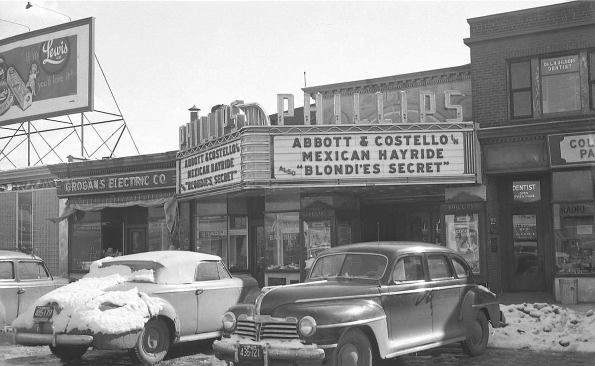Black-and-white photo of a snowy street with vintage cars parked in front of an old movie theater. The marquee advertises "Abbott & Costello Mexican Hayride" and "Blondie's Secret." Other shops and a billboard are visible.