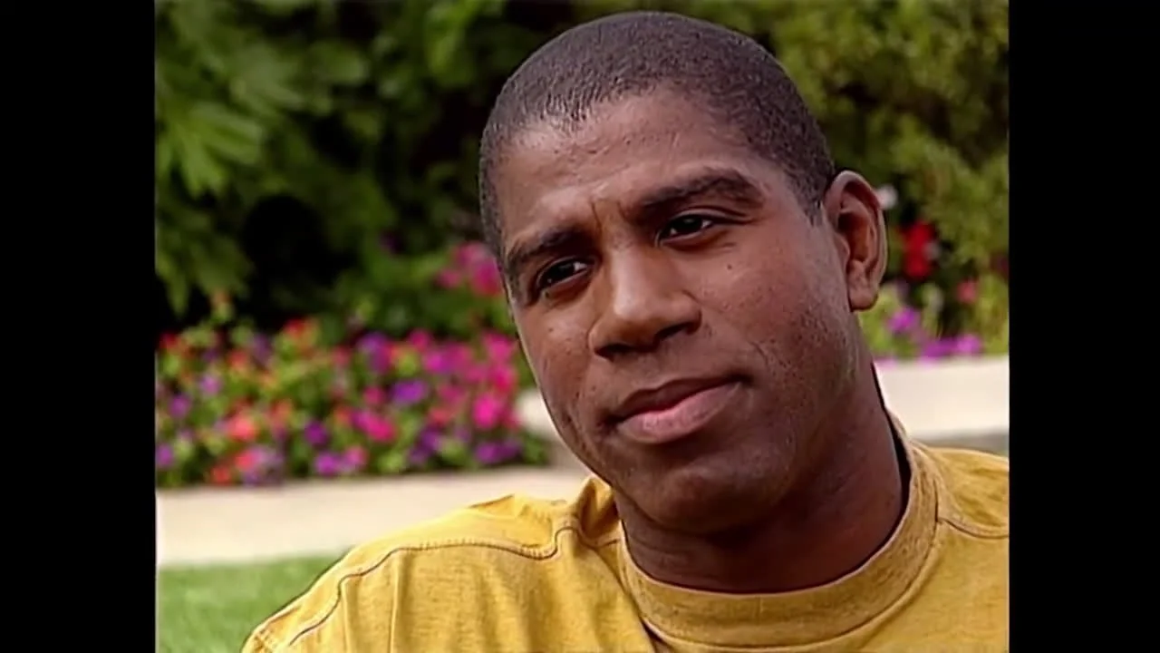 A man wearing a yellow shirt sits outdoors in front of blurred greenery and colorful flowers, looking slightly off camera with a neutral expression.