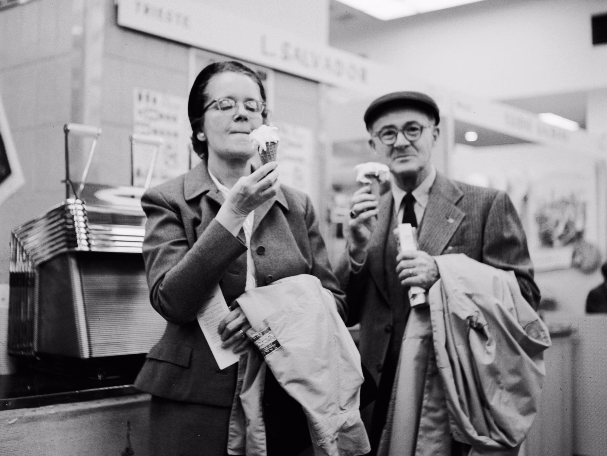 An older woman and man, both wearing coats and glasses, enjoy ice cream cones indoors. They appear happy and are standing close together in what looks like an ice cream shop or cafe.