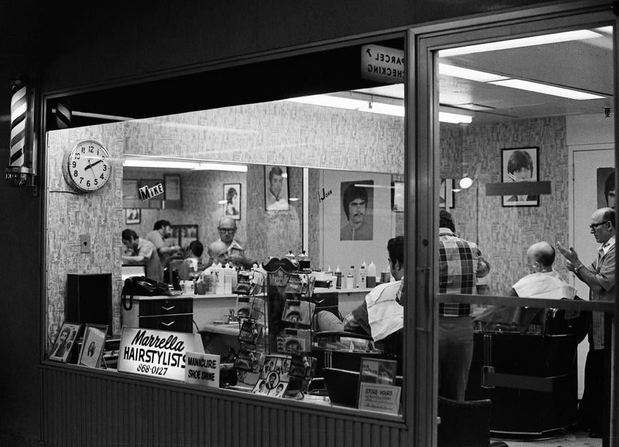 Black-and-white photo of a barbershop with stylists cutting hair. A clock reads 7:06. Barbers and customers are visible; hair products and a "Hairstylist" sign appear in the window. Framed photos hang on the walls.