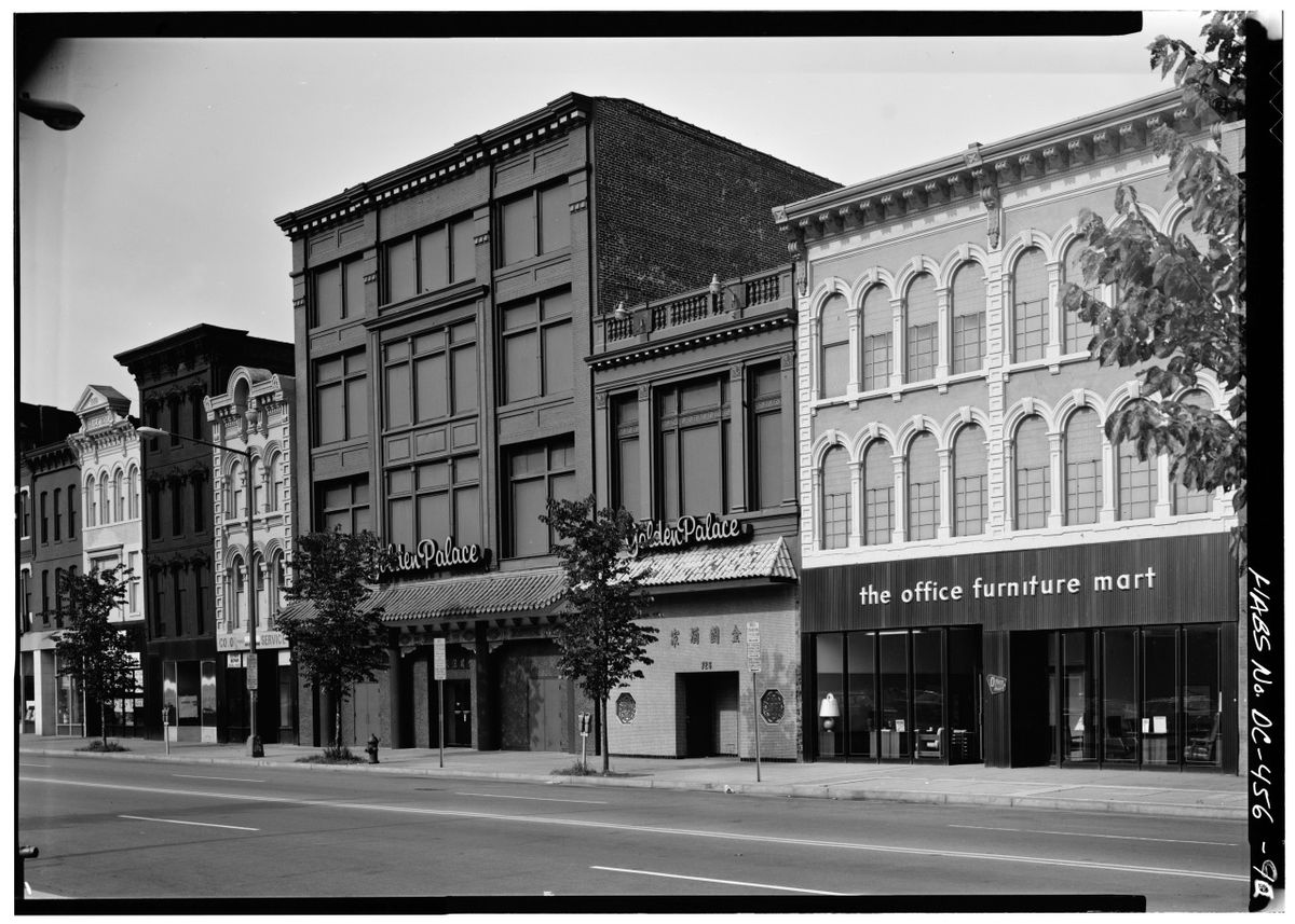 A black-and-white photo of a city street lined with historic commercial buildings, including the Palace theater with its marquee and a storefront labeled "the office furniture mart." The street is quiet with a few small trees.