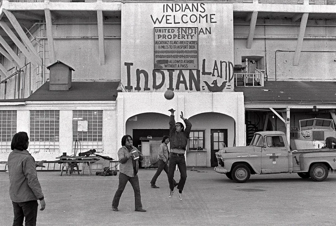 Four people play basketball in front of a building with signs reading "Indians Welcome" and "Indian Land." A truck is parked nearby, and a basketball is mid-air. The scene is from the Alcatraz Island occupation.