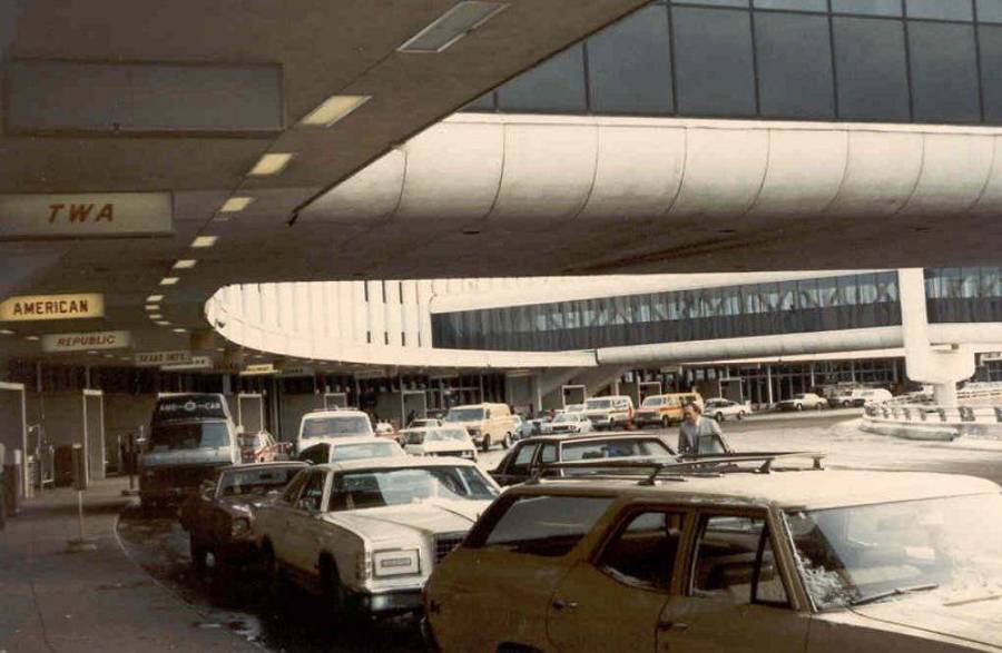 A busy airport curbside from the 1970s or 1980s, with vintage cars lined up, people walking, and signs for TWA, American, and Republic airlines under a curved overhang.