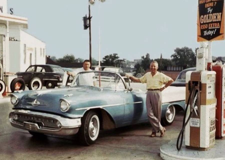 Two men stand by a vintage blue convertible at a gas station. One leans on the car while the other stands by the driver's door. An old gas pump and cars are visible in the background. The scene appears to be from the 1950s.